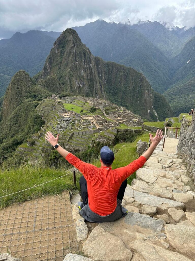Phil Ratcliff seen from behind with arms raised above the ruins of Machu Picchu and Huayna Picchu