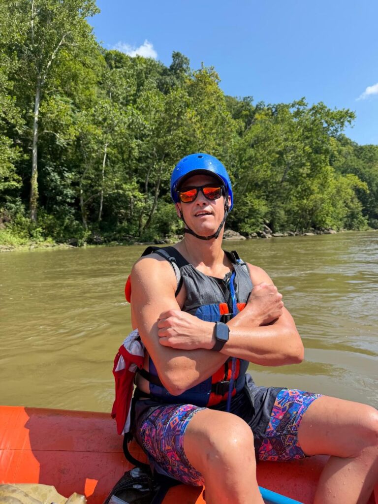Phil Ratcliff in a helmet and life vest with arms crossed on a raft on the New River