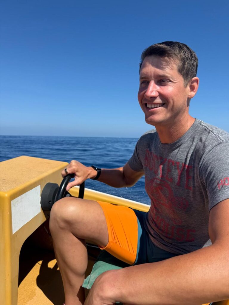 Phil Ratcliff smiling as he steers a small boat on the open Pacific under a clear blue sky